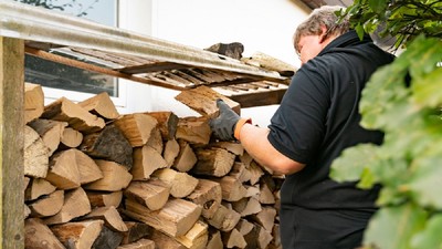 un bărbat, echipat cu mănuși, stivuiește lemnele de foctăiate sub un adăpost din lemn, lângă o casă