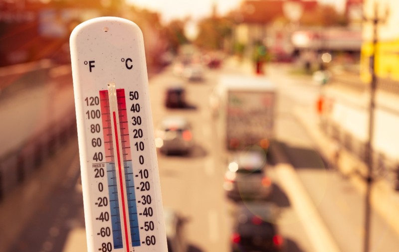 Prim-plan cu un termometru de exterior care indică o temperatură ridicată, pe fundalul unui drum urban însorit și aglomerat cu vehicule.