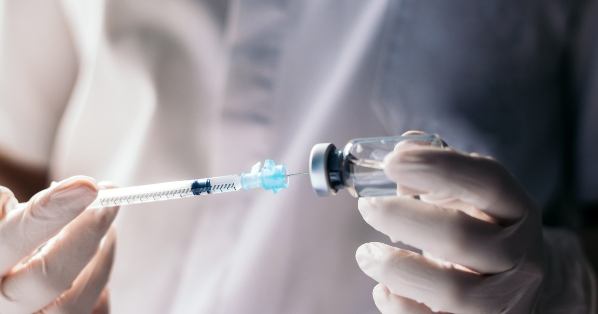 Un cercetător îmbrăcat în echipament medical de protecție pregătește o doză de vaccin, extrăgând lichidul dintr-un flacon cu ajutorul unei seringi. Imaginea sugerează un context de laborator sau de testare clinică, asociat dezvoltării și administrării vaccinurilor.