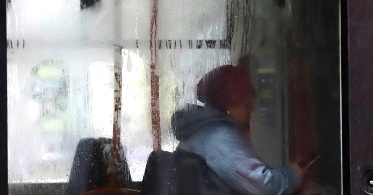 O pensionară este surprinsă din profil, în timp ce stă pe scaun în autobuz, și se uită pe telefon. Femeia este îmbrăcată cu o geacă bleu cu glugă, are căciulă vișine și stă pe scaunul din față al autobuzului, pe al cărui geam se vede condens de la ploaia din ziua respectivă, București, 9 octombrie 2025