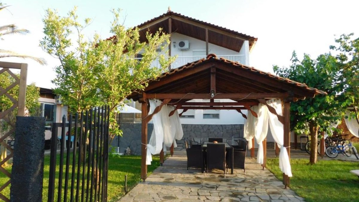 Casa Ferienhaus din Halkidiki, Grecia, cu un foișor în fața casei, iar pe fundal casa de culoare albă și acoperiș de lemn