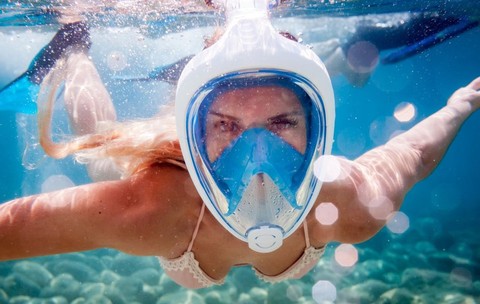 O femeie cu părul blond și ondulat se bucură de o baie răcoritoare sub apă, purtând o mască de snorkeling albă și albastră, care îi acoperă întregul chip