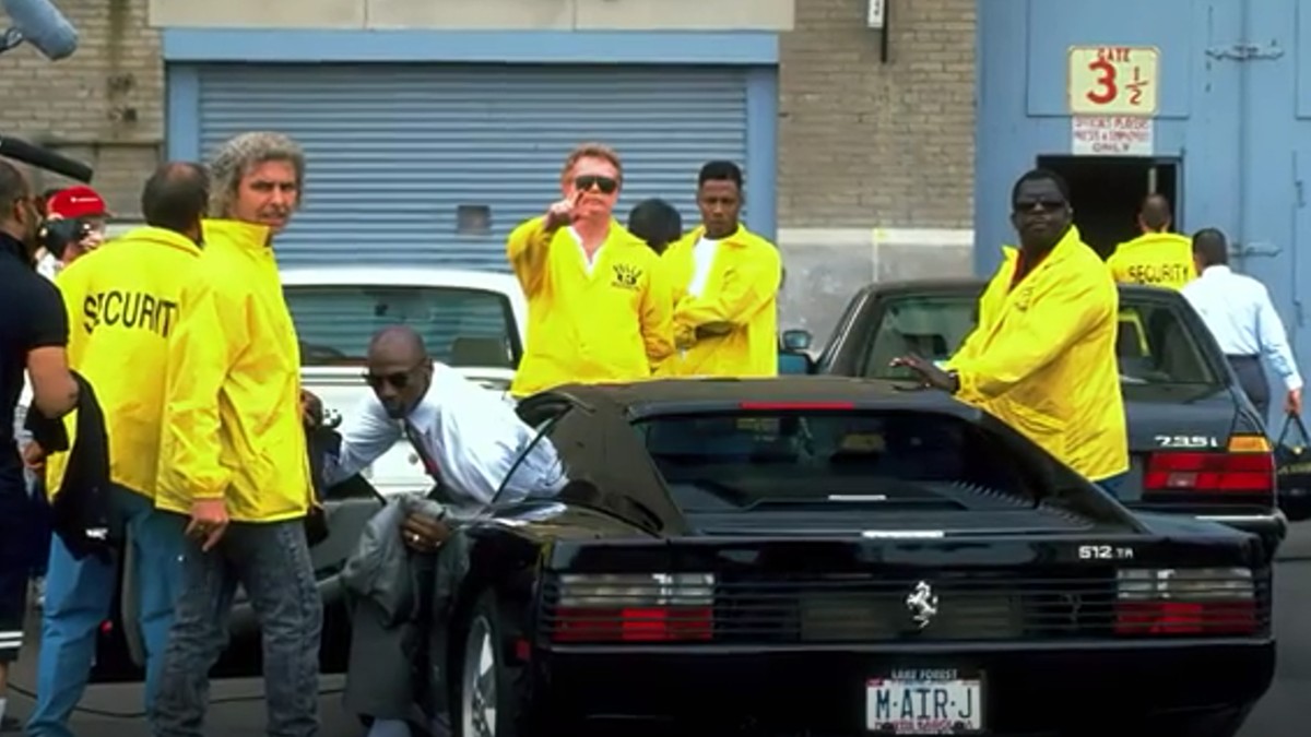 Michael Jordan coboară dintr-un Ferrari negru, înconjurat de agenți de securitate, îmbrăcați în geci galbene.