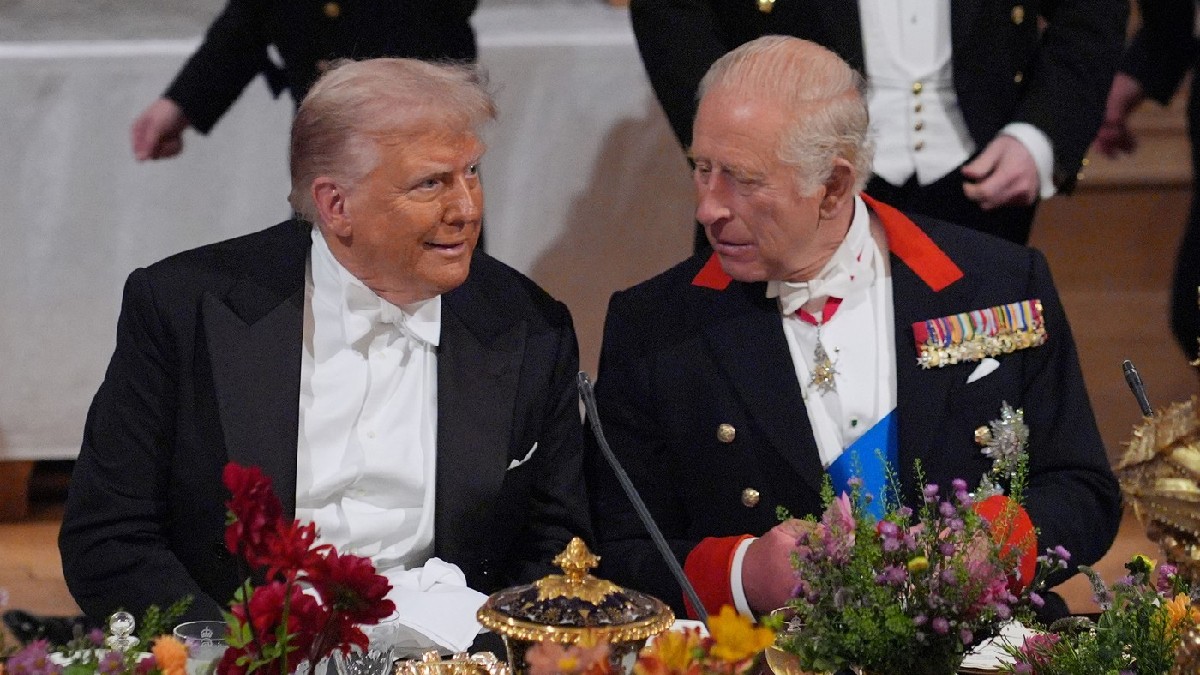 Imaginea surprinde o interacțiune formală între Donald Trump (stânga), îmbrăcat în frac cu papion alb, și Regele Charles al III-lea al Regatului Unit (dreapta), purtând o uniformă militară ceremonială cu medalii și eșarfă. Cei doi bărbați par să poarte o conversație animată la o masă festivă bogat decorată cu flori.