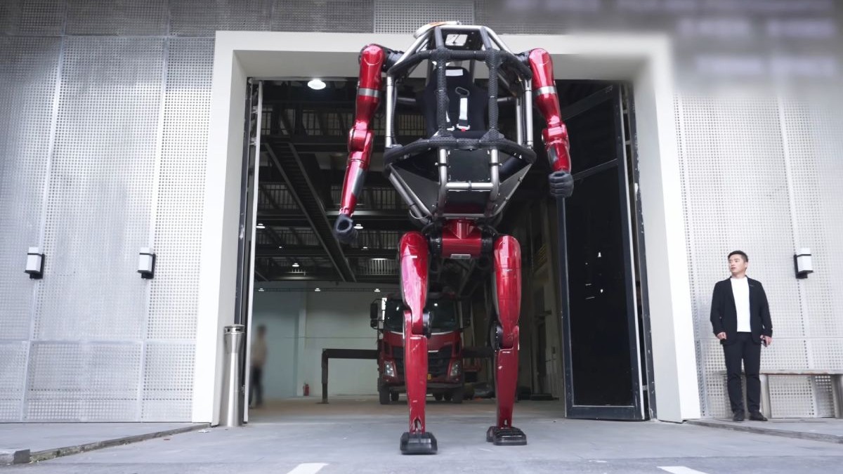 Imaginea prezintă un robot biped de mari dimensiuni, cu o structură metalică roșie și o cabină pentru pilot, ieșind dintr-un garaj industrial în fața unui bărbat care îl privește.