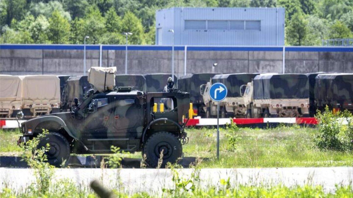 Un vehicul blindat de teren în prim-plan și o serie de camioane militare de transport aliniate în fața unui zid de securitate al unei baze militare.