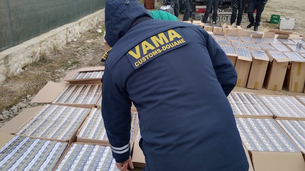 În imagine se observă un lucrător vamal care inspectează mai multe baxuri de țigarete confiscate, dispuse în cutii de carton, în cadrul unei acțiuni de control. Fotografia ilustrează activitatea autorităților vamale de combatere a contrabandei și a traficului ilegal cu produse accizabile.