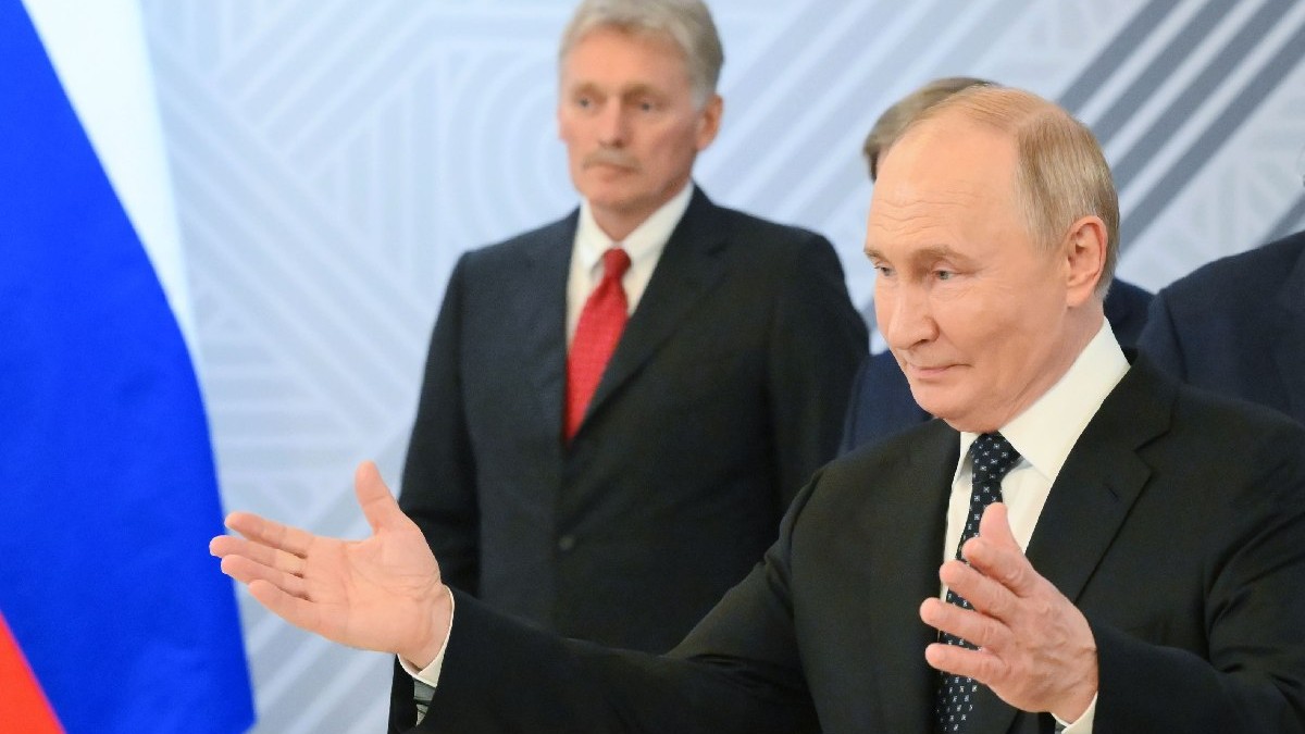 Putin cu brațele deschise, îmbrăcat în costum negru, camasa alba si cravata neagra. În spate, Dmitri Peskov, secretarul de presă al Kremlinului, în costum negru, cîmașă albă și cravată roșie. Președintele rus Vladimir Putin în timpul întâlnirii sale cu președintele egiptean Abdel Fattah El-Sisi în marja celui de-al 16-lea Summit BRICS de la Kazan.