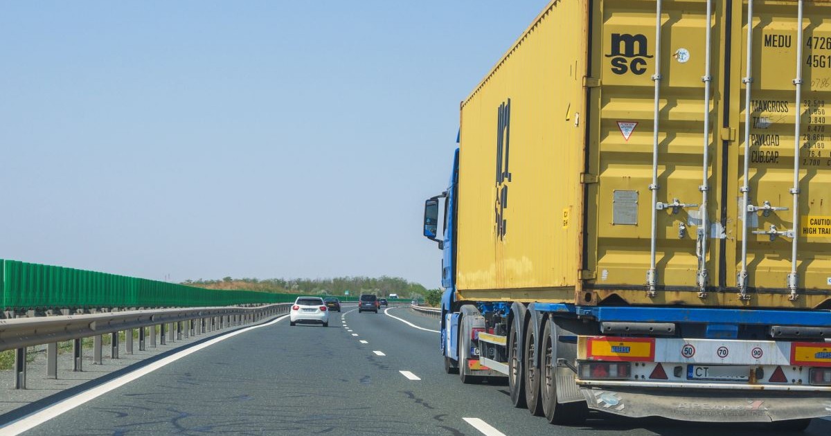 O imagine surprinde traficul de pe o autostradă, având în prim-plan partea din spate a unui autocamion ce transportă un container galben, în timp ce alte autoturisme circulă pe benzile din față sub un cer senin.