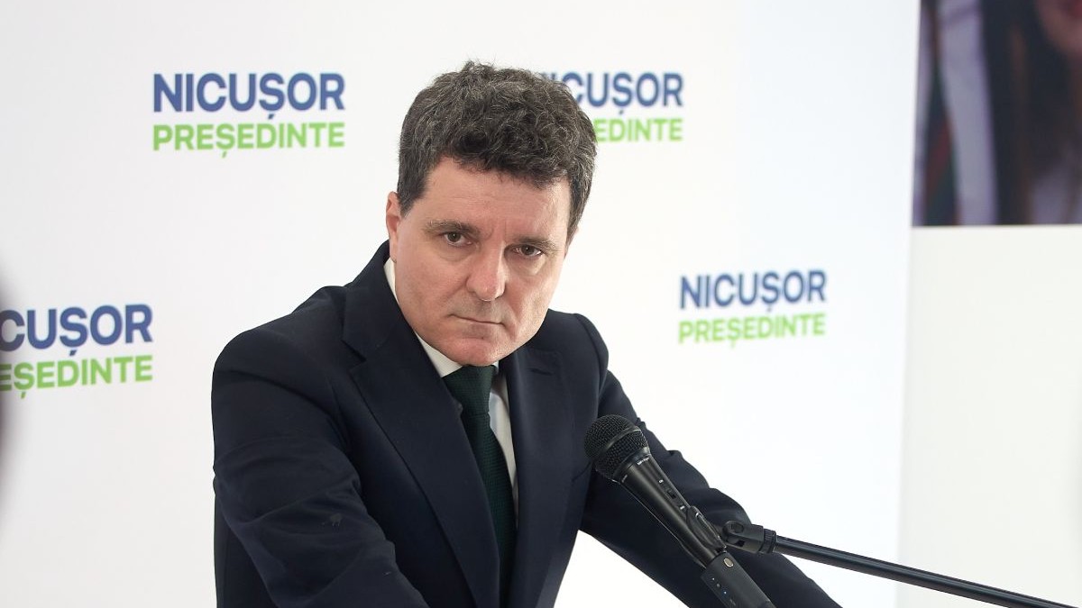 fotografie cu Nicușor Dan in saciu negru, sprijinit cu mainile de un pupitru, cu un microfon in fata, cu un banner alb in spate, pe care scrie Nicusor presedinte