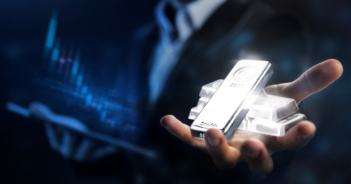 În imagine apare o mână care ține mai multe lingouri de argint, strălucitoare, prezentate într-un mod simbolic și futurist. Fundalul este întunecat, cu nuanțe de albastru, iar în partea stângă se observă graficul unei evoluții financiare, sugerând creșteri pe piețele metalelor prețioase. Silueta unei persoane în costum se distinge în fundal, accentu