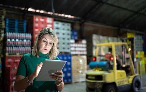 O femeie îmbrăcată într-o bluză verde, utilizând o tabletă digitală într-un depozit industrial cu mărfuri stivuite și un motostivuitor galben în fundal.