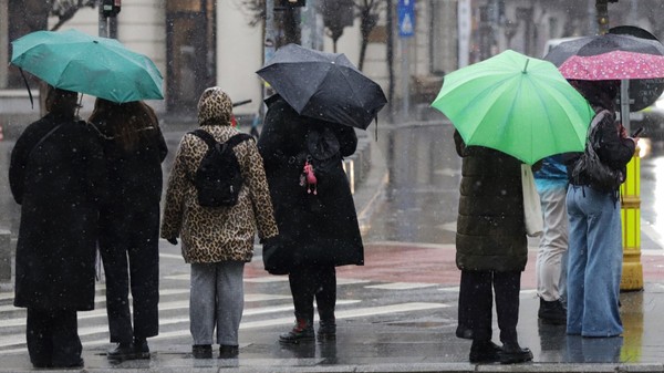 Mai mulți pietoni așteptând la o trecere de pietoni pe o vreme ploioasă și rece, adăpostindu-se sub umbrele colorate și purtând haine groase.