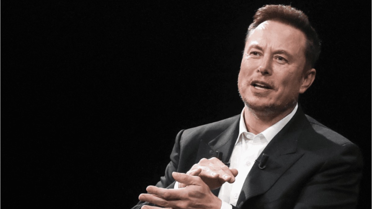 În imagine este ilustrat Elon Musk, în timp ce vorbește. Este îmbrăcat în costum negru, cămașă albă.