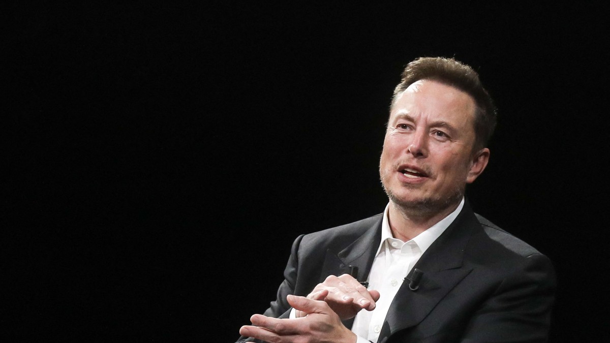Imagine cu Elon Musk pe un Fundal negru, el îmbracat în costum gri închis, in timp ce vorbeste, făcând un semn cu mainile apropiate