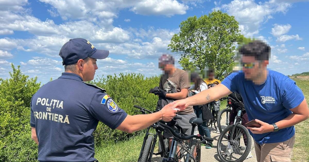 Un polițist de frontieră verifică documentul unui bărbat aflat pe bicicletă, pe un drum îngust în aer liber. În spate se află mai mulți bicicliști care așteaptă, cu fețele blurate. Imaginea este făcută într-o zi însorită, lângă vegetație și copaci, pe traseul de frontieră de pe Canalul Bega.