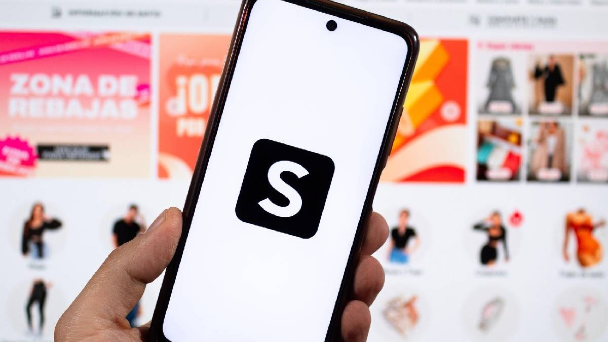O mână masculină ținând un smartphone cu mâna stângă și simbolul Shein și o interfață Shein neclară