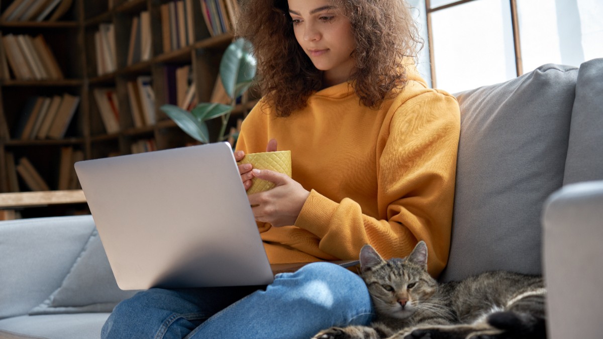 O tânără îmbrăcată în hanorac galben, care stă pe canapea cu o cană în mână și se uită pe laptop