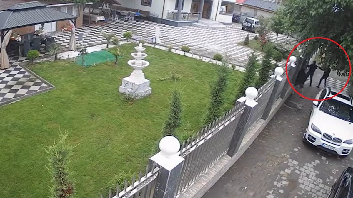 Imaginea surprinde o perspectivă de sus dintr-o cameră de supraveghere asupra unei curți mari cu fântână arteziană, evidențiind într-un cerc roșu mai multe persoane lângă poartă, unde este parcat un SUV alb.