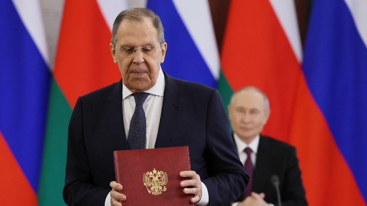 Serghei Lavrov, îmbrăcat în costum negru, cămașă albă și cravată neagră, tine în mână o mapă grena, cu o stemă argintie. În fundal se vede Vloadimir Putin Foto: Hepta