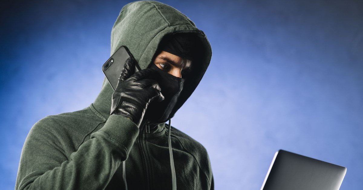 Un tânăr în hanorac verde cu gluga trasă pe cap și cu o cagulă pe față vorbește la telefonul mobil în timp ce se uită pe un laptop