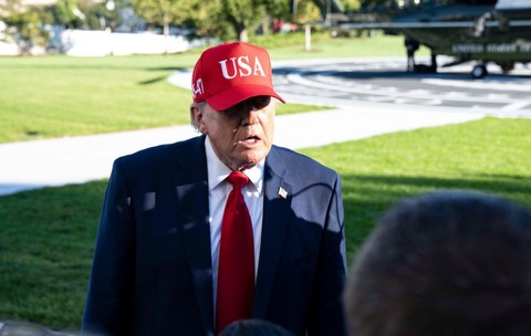 Donald Trump purtând un costum închis la culoare, cravată roșie și o șapcă roșie pe care scrie „USA”, vorbind în aer liber în fața jurnaliștilor, cu un elicopter în fundal.