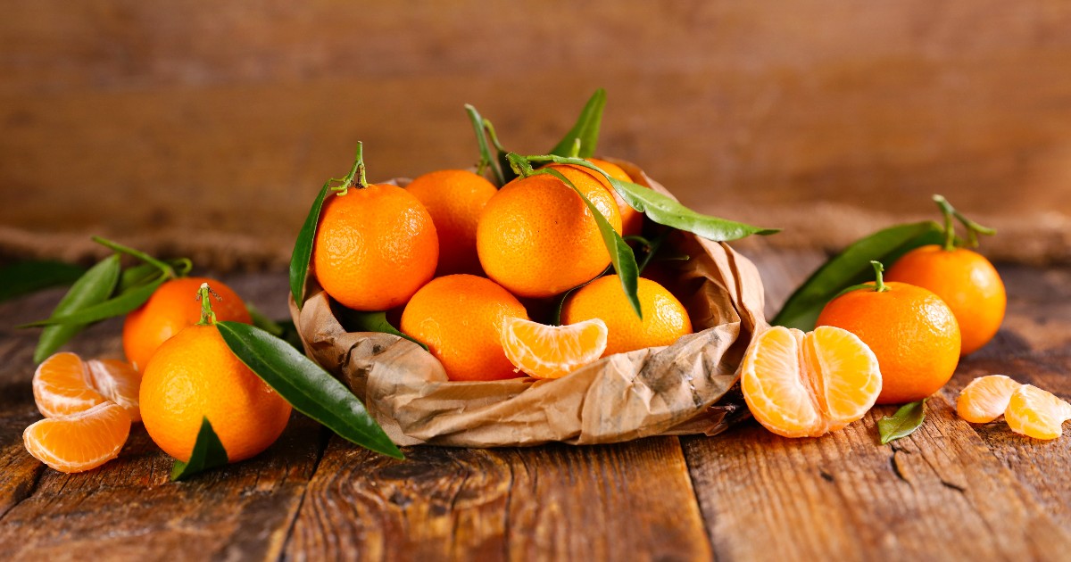 În imagine se văd clementine proaspete, de un portocaliu intens, așezate într-un coș de hârtie pe o masă din lemn. Unele fructe au frunze verzi atașate, iar câteva sunt desfăcute, lăsând să se vadă pulpa suculentă.