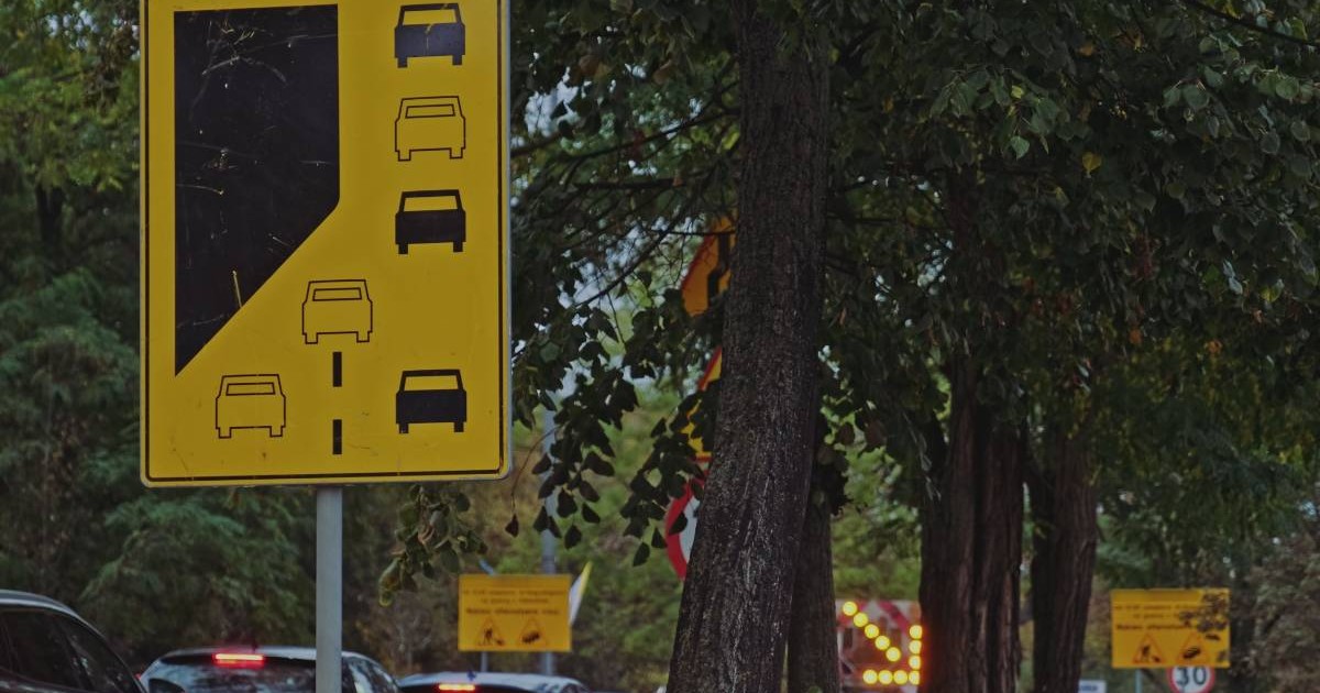 indicator rutier galben care indica regula fermoarului in trafic, pe fundal se observa mai multe masini pe o strada aglomerata din oras. in dreapta sunt niste pomi inalti