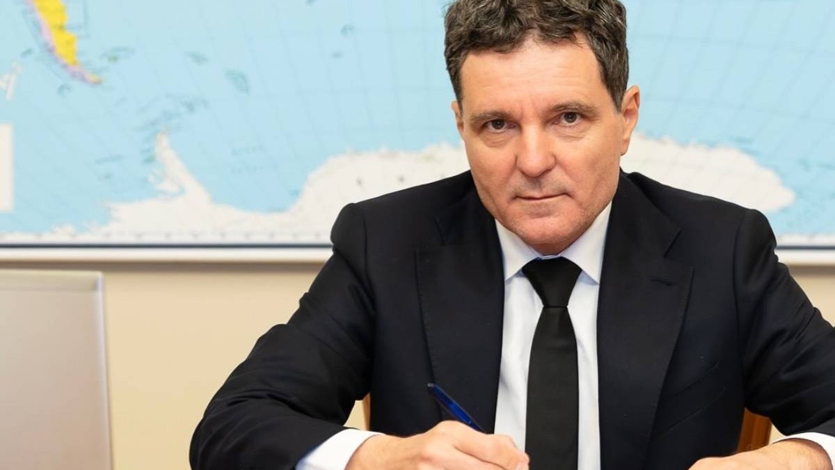 Nicușor Dan, președintele României, în timp ce stă la birou cu pixul în mână, în spatele său are harta lumii și poartă un costum negru