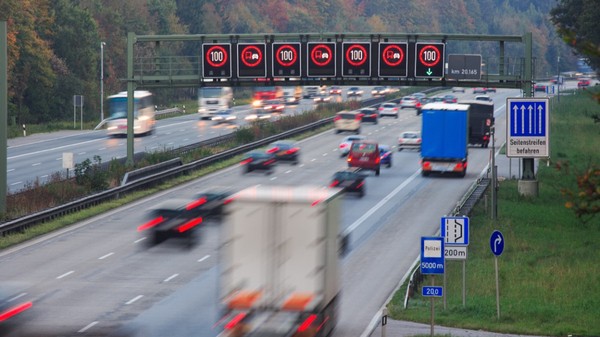 Imaginea prezintă traficul de pe o autostradă din Germania (Autobahn), unde panouri electronice deasupra benzilor afișează o limită de viteză de 100 km/h și restricții de depășire pentru camioane.