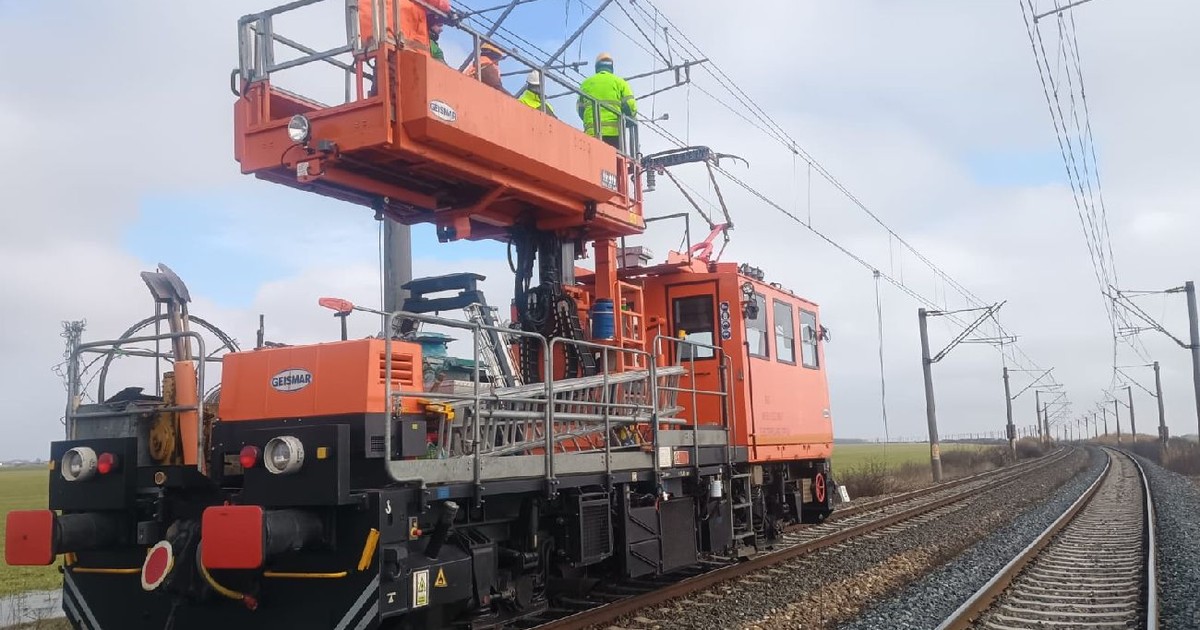 Imaginea surprinde un vehicul de întreținere a trenurilor, pe o linie de cale ferată. Mai mulți muncitori îmbrăcați în veste și căști de protecție de culoare verde neon se află pe platformă.