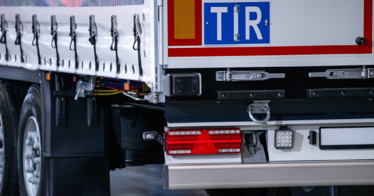 Imaginea prezintă un detaliu din partea posterioară a unei remorci de camion, evidențiind plăcuța albastră cu inscripția „TIR”, sistemul de iluminare și mecanismele de fixare a prelatei.