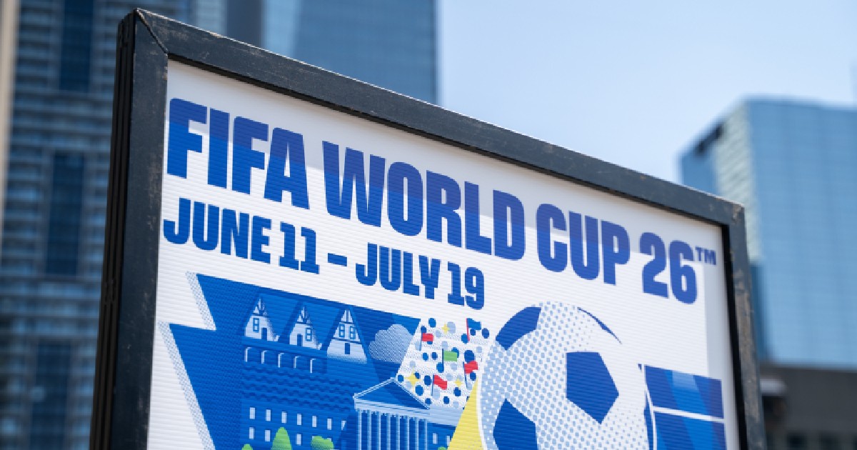 Imaginea prezintă de aproape un panou publicitar care anunță evenimentul sportiv FIFA World Cup '26 (Cupa Mondială 2026), specificând datele de desfășurare: 11 iunie - 19 iulie. Pe panou se află și o reprezentare grafică stilizată, incluzând o minge de fotbal și clădiri, iar în fundal se observă clădiri moderne de birouri.