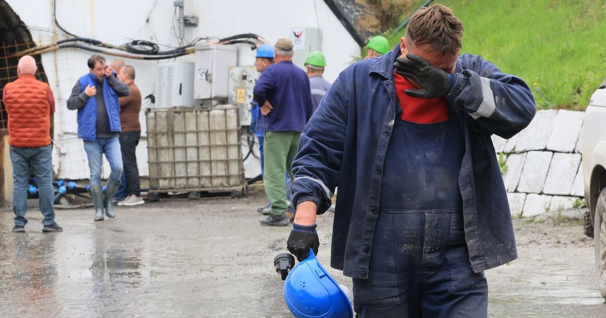 Oameni in uniforma de miner, cu casca si cizme, in fata unei mine, in prim plan un barbat cu mana stanga ridicata la fata si o casca albastra in mana dreapta