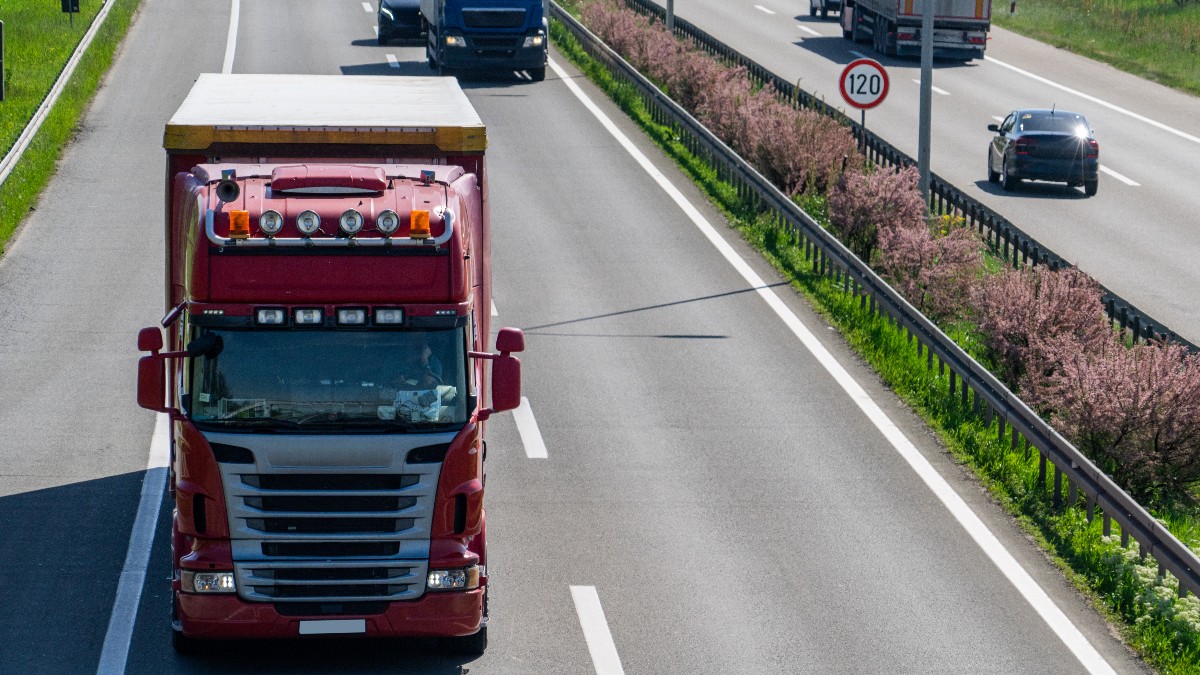 Imaginea înfățișează un camion roșu de mare tonaj care se deplasează pe banda unei autostrăzi, în timp ce pe sensul opus și pe benzile alăturate se observă alte vehicule în trafic.