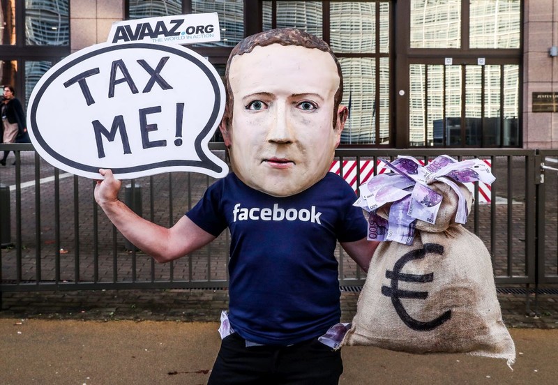 Un protestatar poartă o mască ce îl înfățișează pe CEO-ul Facebook Mark Zuckerberg și ține un banner cu inscripția: „Taxează-mă!”, la o reuniune a miniștrilor de finanțe din UE | FOTO: EPA