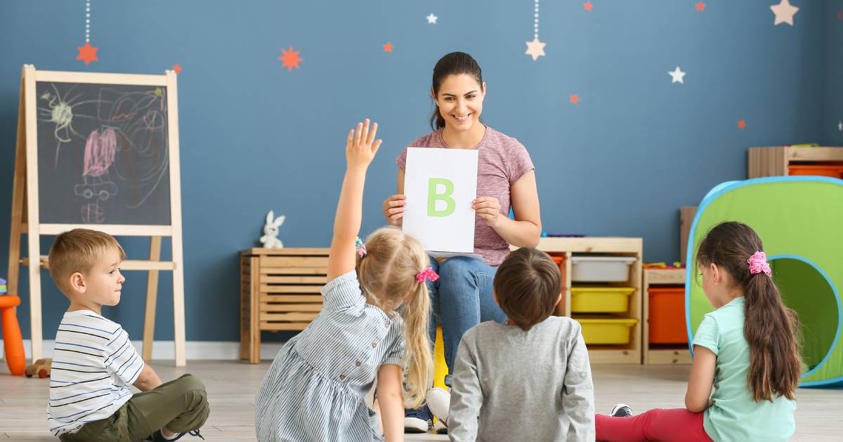 Un grup de copii înscriși deja la creșă sau la grădiniță, în tip ce învață alfabetul de pe o planșă pe care le-o arată educatoarea.