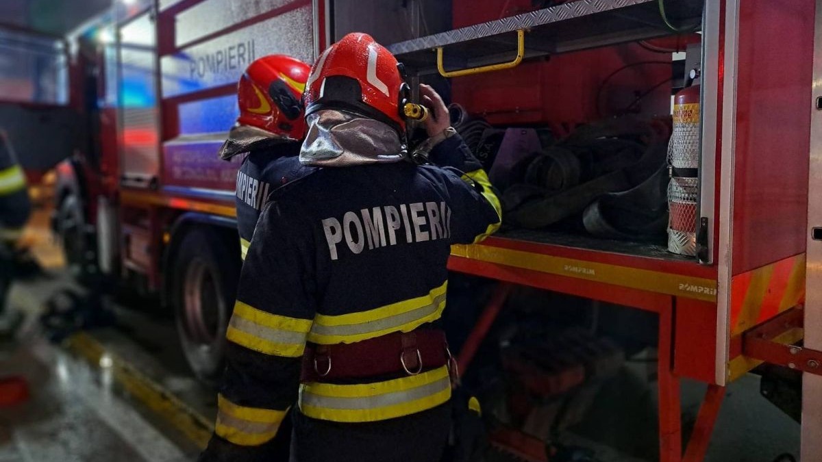 Doi pompieri echipați și cu căști roșii pe cap în prim-plan, fotografiați cu spatele, în timp ce umblă în autospeciala de stingere