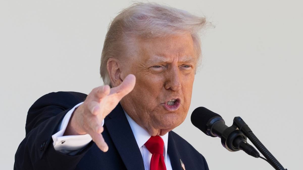 Donald Trump, președintele SUA, in sacou negru, camasă albă si cravata, rosie, cu mana dreapta ridicata si palma deschisa, orientata in fata, surprins in timp ce vorbeste la microfon