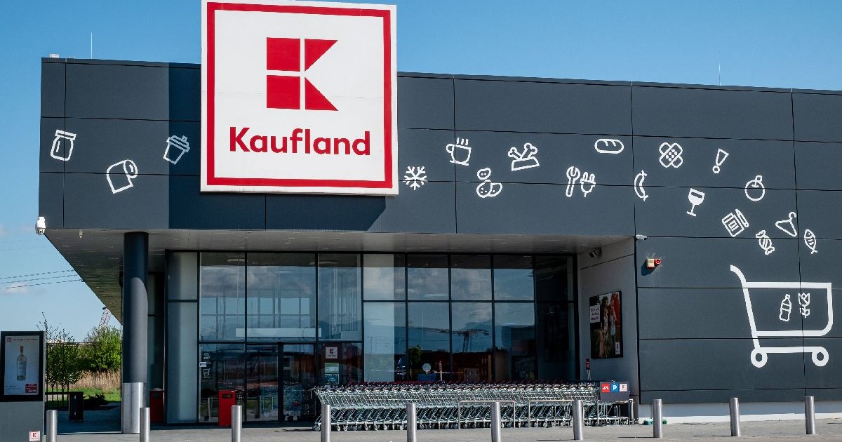 Imaginea arată fațada din sticlă a unui magazin Kaufland, cu sigla la vedere si zeci de cărucioare de cumpărături așezate în rând la intrare.