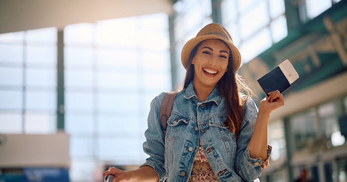O tânără zâmbitoare se află într-un aeroport, ținând în mână un pașaport și un bilet de avion, pregătită de plecare. Imaginea sugerează entuziasmul călătoriilor planificate din timp și ideea de vacanță organizată inteligent.