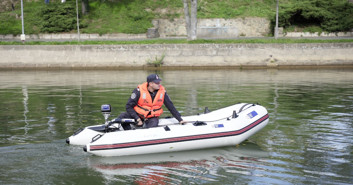 Un pompier într-o barcă, îmbrăcat cu o vestă de salvare portocalie și cu o șapcă pe cap. Barca este pe o apă