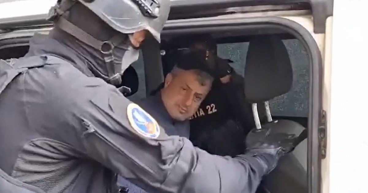 Fotografie cu un polițist în uniformă, cu cască și insignă, care imobilizează un bărbat cu părul scurt în interiorul unui vehicul.