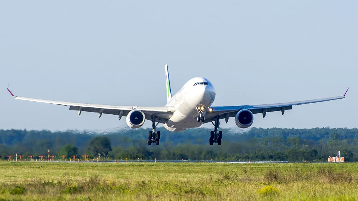 Imaginea surprinde un avion de mari dimensiuni în momentul decolării sau aterizării, cu trenul de aterizare scos, deasupra unei piste mărginite de vegetație.