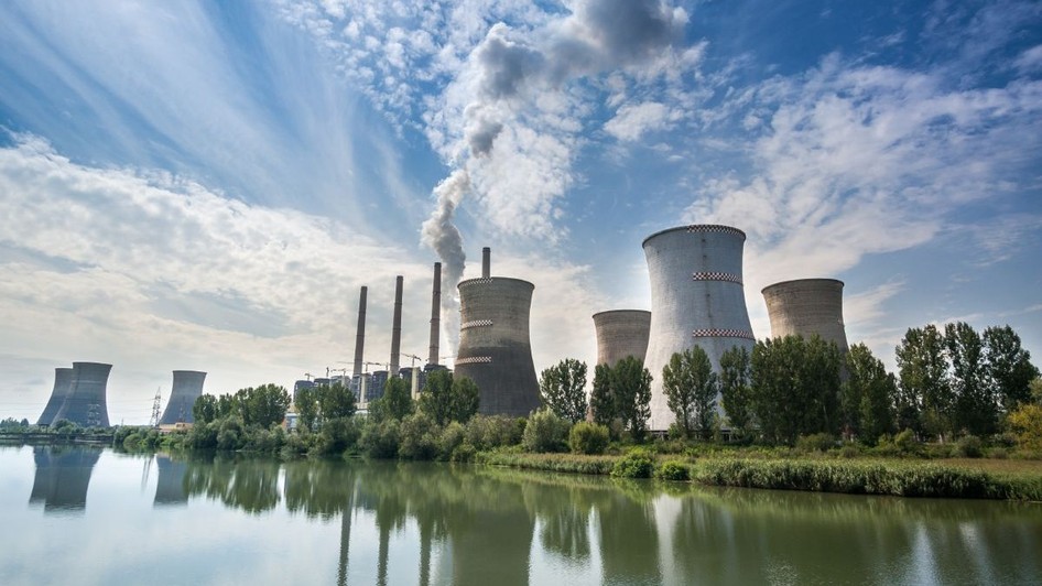 Termocentrala Turceni, cu un lac în față, turnurile uriașe pe fundal pe care iese fum și cerul albastru cu nori albi