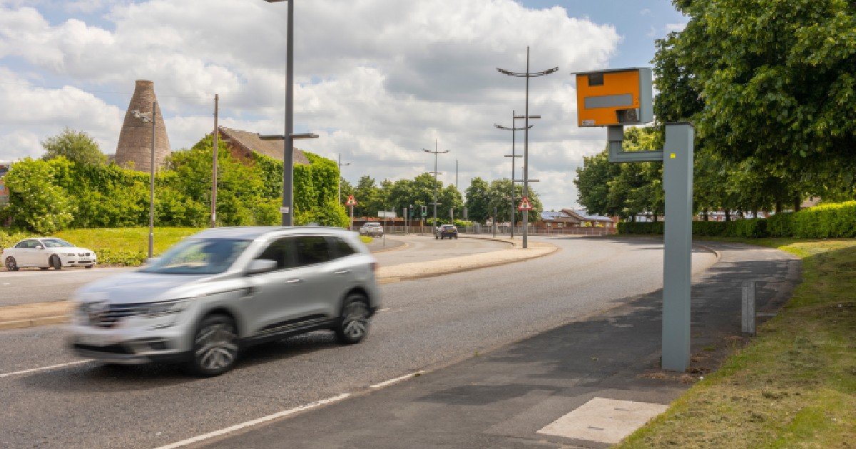 Imaginea prezintă un autoturism argintiu în mișcare pe o șosea, monitorizată de o cameră radar portocalie amplasată pe marginea drumului, într-o zonă cu vegetație și clădiri în fundal.