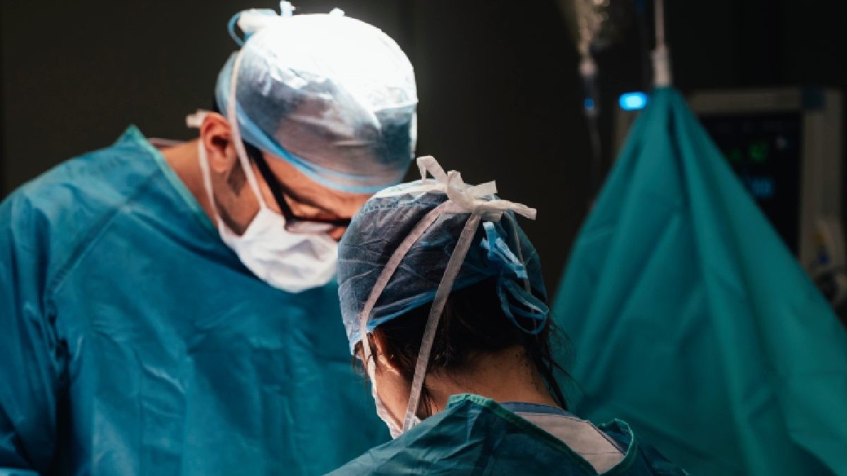 Doi medici, costumati in halate cu bonete si masti, care opereaza in sala de operatie