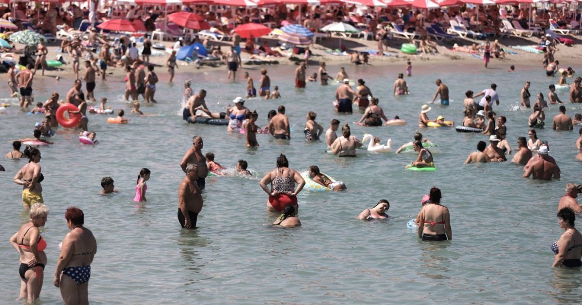 Mai mulți oameni în costume de baie sunt în apa mării, se văd și copii cu colace, iar în spate sunt umbrele mari de soare, roșii, unde turiștii stau la umbră