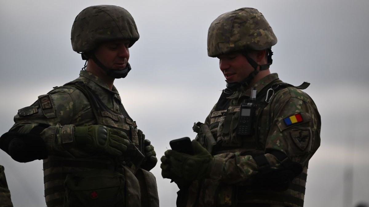 Doi militari români, îmbrăcați în uniforme, cu căști pe cap, cu cerul înnorat pe fundal