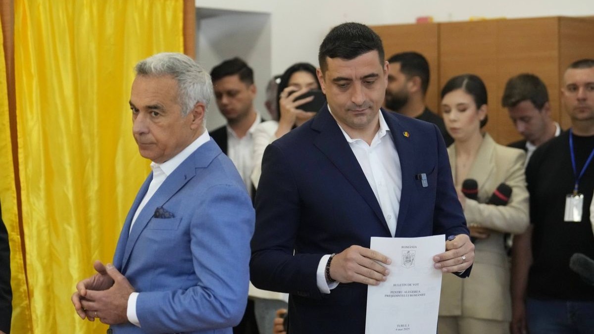 In Imagine George Simion, stanga, în costum bleu și cămașă albă, alături de George Simion, dreapta, in sacou negru si cămasp albă, cu buletinul de vot în mână, stau spate în spate in fata urnei, in secțoa de votare